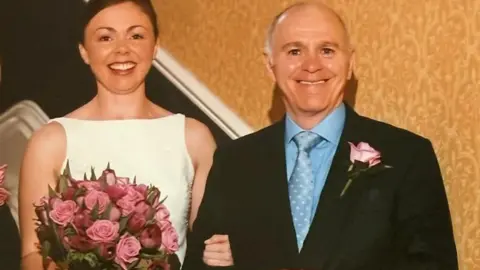 Tara Ingle A beaming bride in a white wedding dress holding a bunch of roses and linking arms with a smiling gentleman in a smart dark suit.