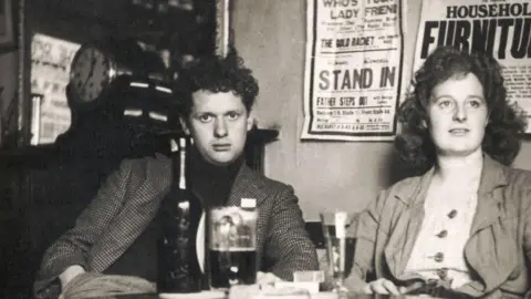 Getty Images Dylan Thomas, pictured with his wife Caitlin, sitting down at a pub table with pints of beer. He is wearing a chequered jacket and looking straight at the camera. Caitlin is looking to the side, her curly hair is loose and she is wearing a jacket with a buttoned top underneath. 