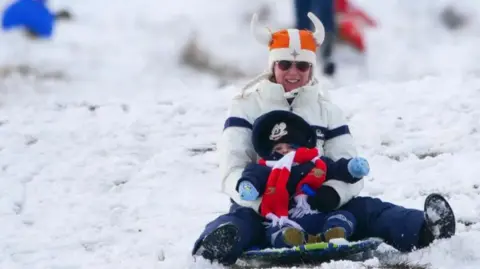 PA Media Mother and toddler sledging downhill in snow wearing hats and woman wearing sunglasses and weatherproof trousers and snow boots