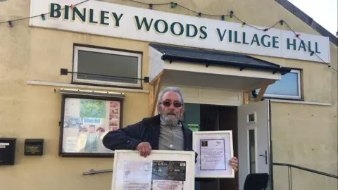 Norman Miller stood outside Binley Woods village hall. He has slicked back grey hair and a grey beard, he is wearing dark sunglasses and a grey quater zip jumper underneath a navy blue jacket. He is holding two picture frames.