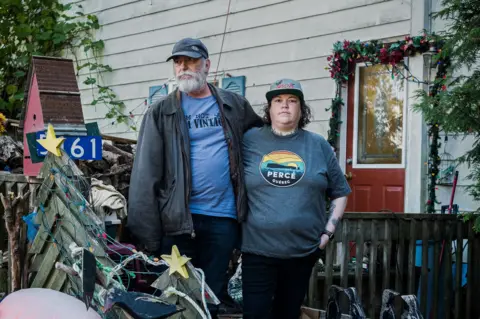 Derek Cuthbertson and his step-daughter Jillian Lucas, both in T-shirts and baseball caps, stand close together outside his white house with a red door, surrounded by fishing buoys and trees.