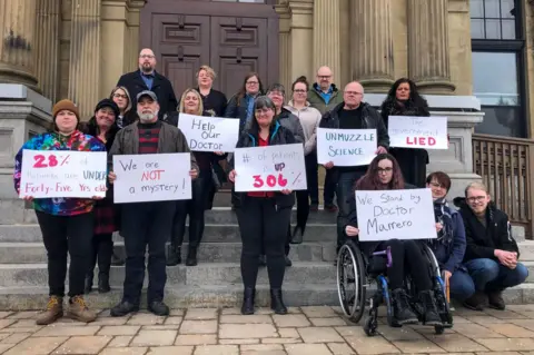 Global News Jillian Lucas and other patients stand in front of a government building holding signs saying 'We are not a mystery, 'We stand by Doctor Marrero' and other slogans. 
