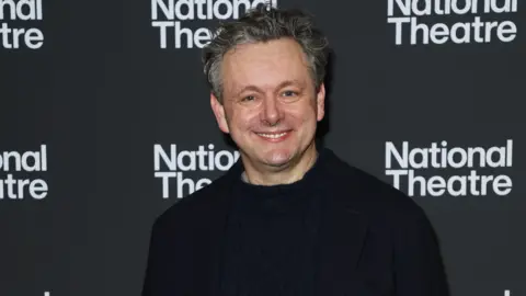 Getty Images Michael Sheen posing and smiling in front of a National Theatre-branded backdrop