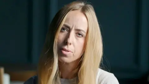 BBC reporter Catrin Nye looks ahead with her head tilted. She has long blonde hair and is wearing a grey top. There is a dark coloured background behind her.