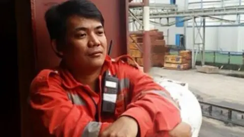 PA Media Solong crew member Mark Angelo Pernia. He is wearing red overhauls and has a black walkie talkie attached to his collar. He has short dark hair in a parting and looking out beyond the camera.