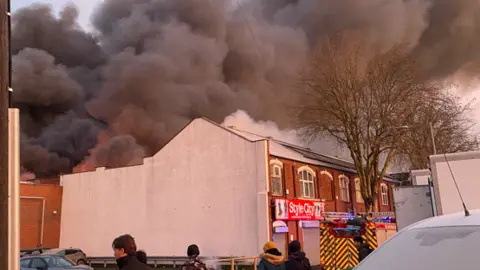 People stand in a street near to the fire as dark smoke plumes billow in the sky over a row of neighbouring shops. A fire engine can be seen parked in the street by some shops. 