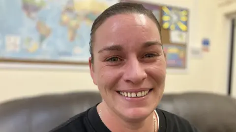 Holly has dark hair tied back and brown eyes. She wears a black top. She is giving a big smile for the camera.