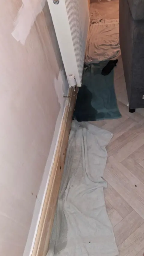 Jane Wallbank Towels are placed across a floor under a radiator and skirting board.  The towels are clearly wet soaking up water from the 