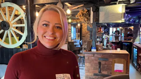 Nicki Bird, who has short blonde hair and is wearing a dark red turtleneck jumper, stands and smiles at the camera. She is in a pub and the bar is in the background.