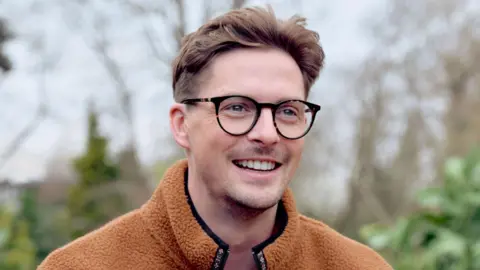 Olivia Spencer Alex George wearing a fleece and glasses smiles. It is a head and shoulders shot.