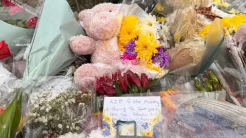 Jasmine Lowe / BBC A close-up shot of flowers. In the centre is a pink teddy bear above a note that reads: RIP to the mummy trying to save your little girl.