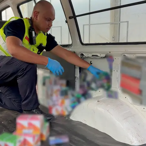 Singapore Immigration and Checkpoints Authority A Singaporean customs officer pulls out boxes of vaping supplies from false compartments inside a van. The boxes have been blurred out by authorities.