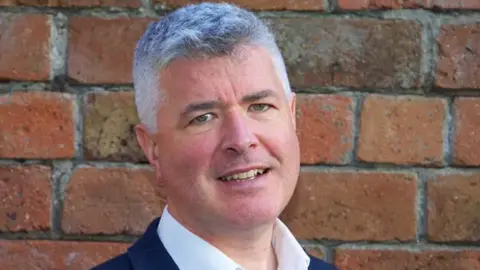 Scottish Information Commissioner Man with short grey hair wearing a white shirt and dark jacket, standing in front of a red brick wall and smiling slightly at the camera.