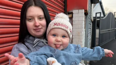 Close up of Alex holding Lilah in front of a red garage. Lilah has a big smile and is looking at the camera with her tongue out. 