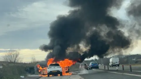 Alex McClean A white car engulfed in orange flames with a large black cloud of smoke rising in the air. The car is parked in a lay-by while other cars are still driving on the road.