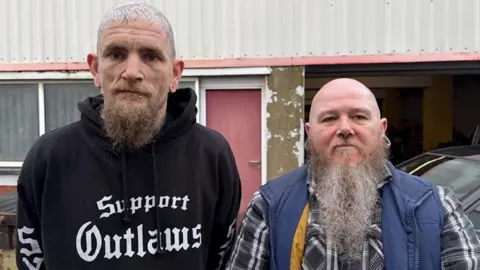 Wesley and Marc pictured standing next to each other in front of a metal warehouse unit. They are both looking at the camera with a serious look. 