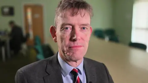 Paul Moseley/BBC Jonathan Hall, Hellesdon Parish Council's clerk looks to camera. He has whispy light brown hair and is in a dark suit, light blue shirt and has a red, blue and gold striped tie on. 