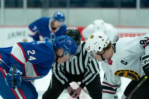 Sabrina Lantos/HBO Max Two men from rival teams face each other on an ice hockey rink. On the left the man is in a blue and on the right the man is dressed in white. There is a referee in a black and white striped uniform in between them