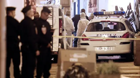 AFP French policemen and forensics investigate around a crime scene - with police in dark uniforms unfocused in the foreground and a white police car and forensics officers in white suits in the background