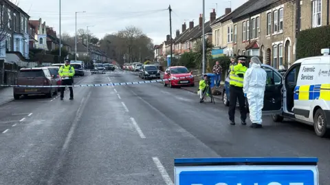 Richard Knights/BBC A police officer is standing in the road behind a cordon. Another officer is crouching behind a camera on a tripod and a second uniformed officer is speaking to a forensic investigator, dressed in white overalls, and standing next to a forensic services van. Parked cars are inside the cordon and terraced houses are on either side of the street.