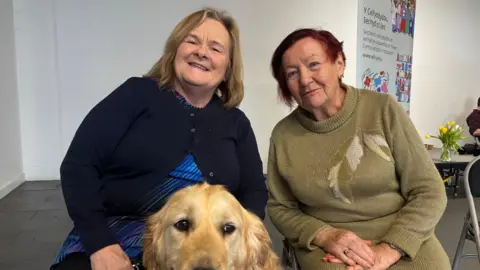 Two older women are sitting next to each other looking at the camera. The one on the left has blonde hair and is wearing a navy cardigan. In front of her is her golden retriever guide dog. Sitting on the right is a lady with short, red hair and she is wearing a long, green jumper.
