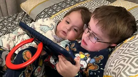 Cartmill family A young boy and girl and lying on a bed reading a tablet.