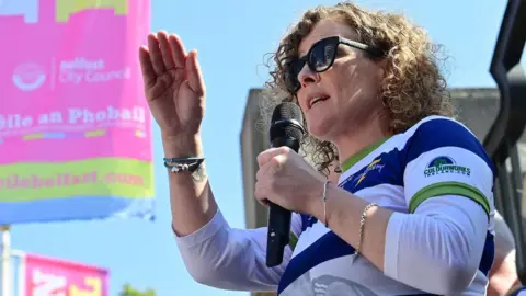 Pacemaker Fiona Donohoe has curly blond hair and is wearing a blue and white striped sports top with her son's image on it and sunglasses as she addresses a crowd with a microphone