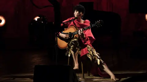 BBC Studios Jacob is sitting on a stool playing a guitar and singing into a microphone. They are performing barefoot in a pair of golden-feathered trousers and a red shawl.