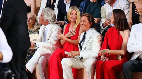 Getty Images Giancarlo Giammetti, Gwyneth Paltrow and Valentino Garavani attend the Valentino Mirabilia Romae Fashion show at Piazza Mignanelli on July 9, 2015 in Rome, Italy. Giancarlo and Valentino are sitting either side of Paltrow, both wearing white suits. Paltrow, in the middle is smiling, holding one hand to her chest, with Valentino sitting next to her in the front row of the show smiling