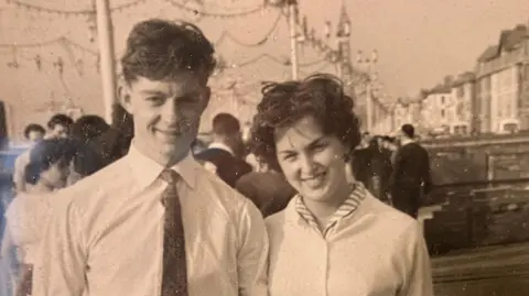 Family handout A black and white photo of Dave and Pam Brazier, a young man in a white shirt and tie and a woman in a light-coloured cardigan are stood together in a busy street and smiling at the camera.