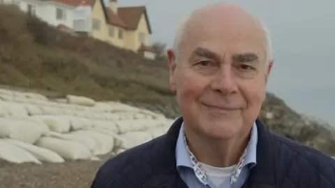 Jamie Niblock/BBC Mark Packard smiles at the camera on a beach that is covered in white sandbags as part of sea defences. He has some grey hair. He wears a navy coat, a blue shirt and a white T-shirt underneath. There are homes on the cliff behind him in the background.