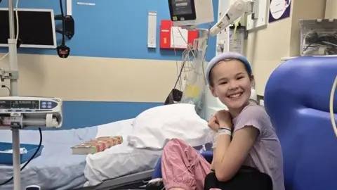Diabetes UK Imogen sits in a hospital chair, next to a bed, with a big smile on her face. She is receiving immunotherapy via an infusion into a vein. The infusion pump machine is visible to her right.