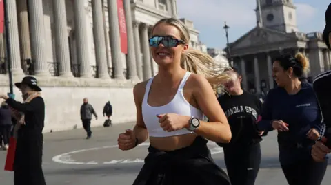 Maddie Albon Maddie, who has long blond hair tied back, is on a run with other people. She wears a white top, smart watch and sunglasses - there are buildings in the background. 