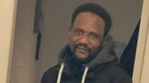 Facebook An African man with short black hair and a small black beard, smiles at the camera. He is wearing a dark hoodie and jacket.