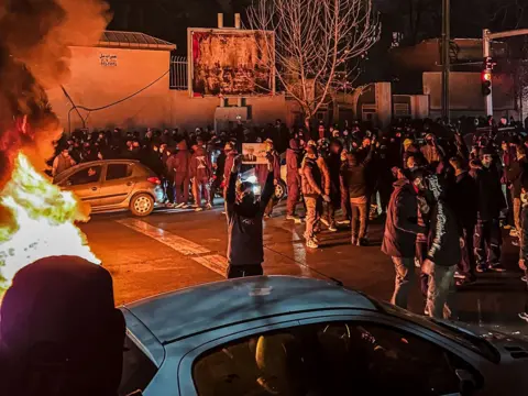 Getty Images Iranians gather while blocking a street during a protest in Tehran, Iran on 9 January. One protester wearing a face covering stands in the centre of the road holding up an image while a fire burns 