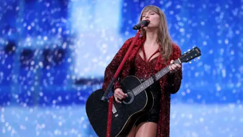 Getty Images Taylor Swift performing on stage. She is playing a black guitar and singing in to a microphone. She is wearing a red sparkly outfit with a red sparkly jacket. 