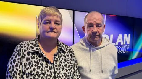 Linda has short blonde hair and is wearing a leopard print shirt. George has very short hair and is wearing a grey hoodie. They are standing in front of TV's which say 'The Nolan Show'.