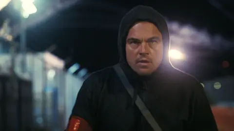 Warner Bros A still from One Battle After Another showing Leonardo DiCaprio's character wearing a black hoodie and walking intently at night