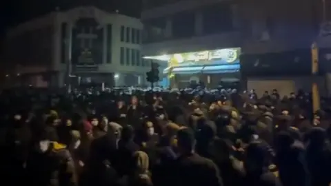 Phone footage Screenshot from phone footage showing large crowd of people gathered outside after nightfall, with light from a building illuminating the dark