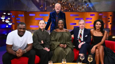 PA Media The five Graham Norton Show guests sat on the famous red sofa, with a table covered in drinks glasses in front of them. Behind them stands Graham Norton in a suit with grey hair. Behind the whole group is a brightly-coloured television studio.