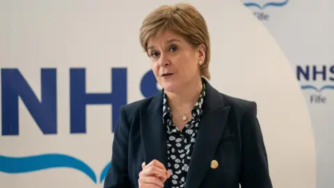 Reuters Nicola Sturgeon standing at a lectern in front of a backdrop branded with NHS Fife's logo. She is wearing a blue blazer with gold buttons and a navy and white patterned blouse.