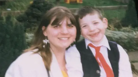 Monica Kershaw pictured with her son - it's an old photo, with Christopher looking to be very young, possible 10 to 12 years old. He is smiling widely and wearing a red tie and waistcoat, next to his mother who is also smiling and wearing an off white jacket. 