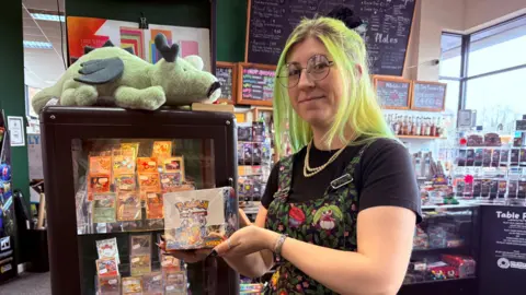 Amy Wosley Amy standing in a café surrounded by trading cards holding a Pokémon box