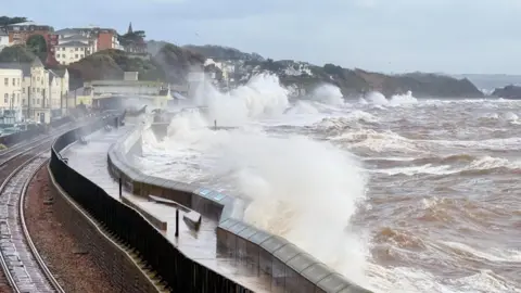 Weather Watchers/AstroPostie The picture shows huge waves crashing over a sea wall during rough weather. Brown, churning water is surging in from the right, and the force of the waves is sending spray high into the air. 