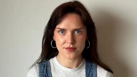 Keelin Moncrieff Keelin stares into the camera. She has blue eyes and silver hoop earrings, wearing her brunette hair down. She is wearing a grey t-shirt and we can see the shoulder straps of her dungarees. She stands against a plain white wall.