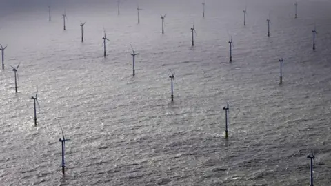 AFP via Getty Images An aerial image of 3/4 rows of wind turbines in the middle of the ocean forming a wind farm in the North Sea. Each turbine has a blade at the top and is supported by a pole above water and a base under water that is not visible in the photo. 