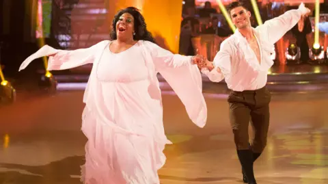 Alison Hammond on a ballroom dance floor performing with partner Aljaz Skorjanec. She is wearing a long pink flowing dress and he black trousers and a ruffled white shirt. He is holding her hand and both have their arms open wide and are smiling broadly.