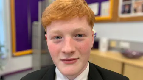 Oliver has red hair and light blue eyes. He is wearing a black blazer and white shirt.