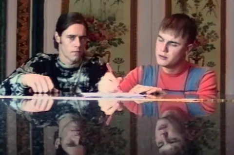 Netflix Howard Donald and Gary Barlow handwrite lyrics while sitting at a piano. Their faces are reflected in the piano lid.
