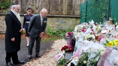 PA Media King Charles in a grey suit looks at flowers and tributes left outside the synagogue. Rabbi Daniel Walker is standing to his left.